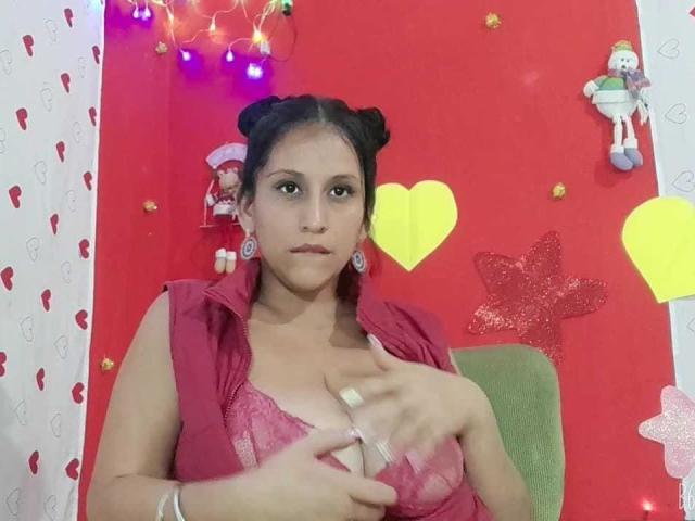 KattyBoobsLove, avec des seins de taille moyenne, portant un soutien-gorge en dentelle rose et un gilet assorti, tenant ses seins, cheveux noirs en deux chignons, boucles d'oreilles rouges, chaise verte et décorations murales colorées..