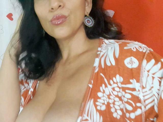 KattyBoobsLove met glanzende, roze lippen, donker, golvend haar en een oranje top met bloemen, die haar grote, onbedekte borsten onthult. Ze draagt een ronde oorbellen en ziet er schattig uit..