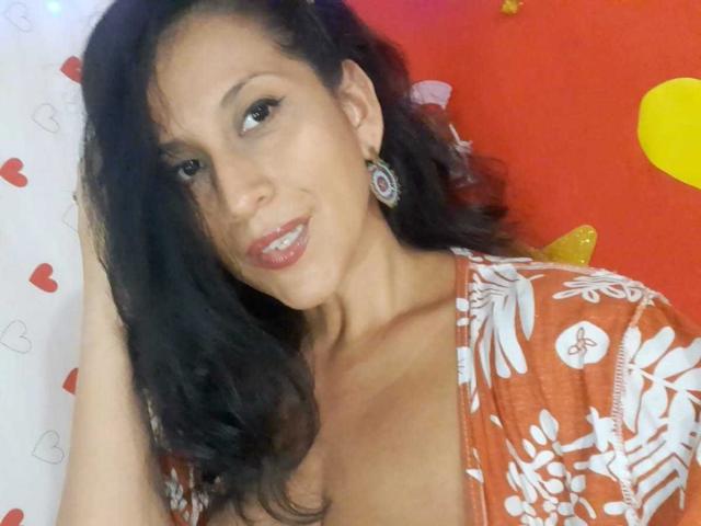 KattyBoobsLove, avec ses cheveux noirs ondulés, sa peau brune et son sourire charmant, porte un haut orange à motifs floraux, laissant entrevoir un léger décolleté, et des boucles d'oreilles argentées, tout en regardant l'appareil photo..