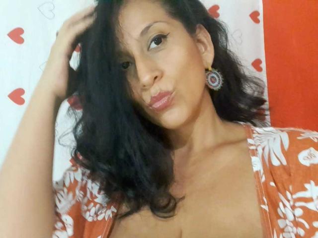 KattyBoobsLove tem cabelos castanhos ondulados, pele morena e está vestindo uma blusa laranja com estampas florais, que mostra o colo, com uma mão no cabelo, e usando um brinco em formato de coração..