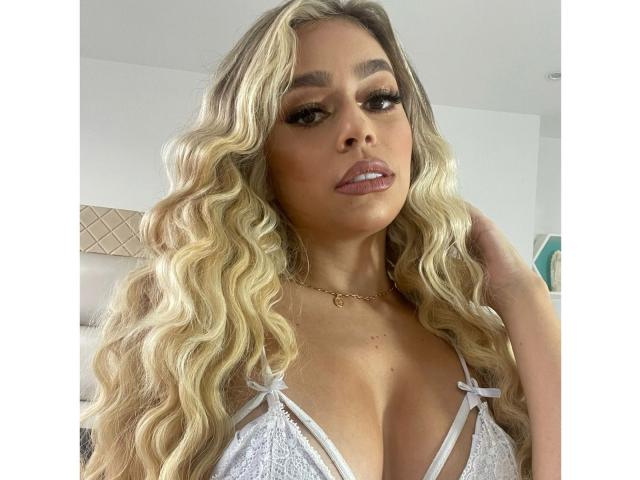 Julietes a de longs cheveux blonds ondulés, des sourcils épais, et elle porte un soutien-gorge en dentelle blanche qui révèle ses seins gros et ronds. Elle regarde l'appareil photo avec un léger sourire..