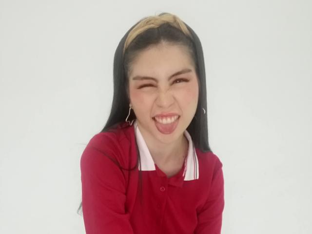 IsaTimeSweet with black hair and tan streak, winking and smiling with tongue out, wearing a red polo shirt with white collar, against a white background.