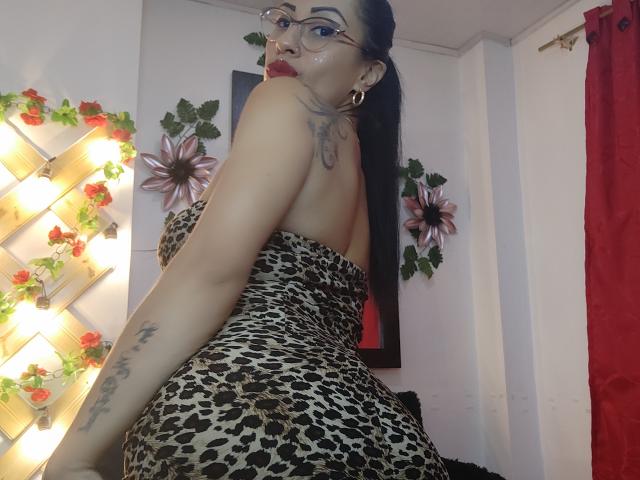 LunaCandy con gli occhiali, capelli scuri raccolti in una coda di cavallo, indossa un abito a macchie di leopardo senza spalline, mostrando la schiena e il braccio tatuato, guarda oltre la spalla, rossetto rosso, orecchini a cerchio..