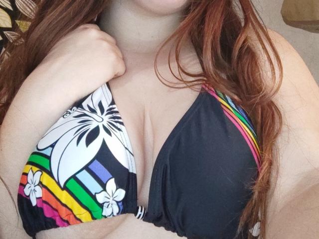 Les seins volumineux et crème de SexMorey sont à peine couverts par un haut de bikini coloré à motifs floraux, une main agrippant le tissu, mettant en valeur ses cheveux roux et ondulés..