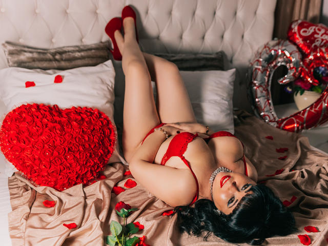 MatureVivian lying on a bed in red lingerie, her hands on her pussy, wearing red heels, with rose petals and a heart-shaped red rose pillow. She's looking at the camera.