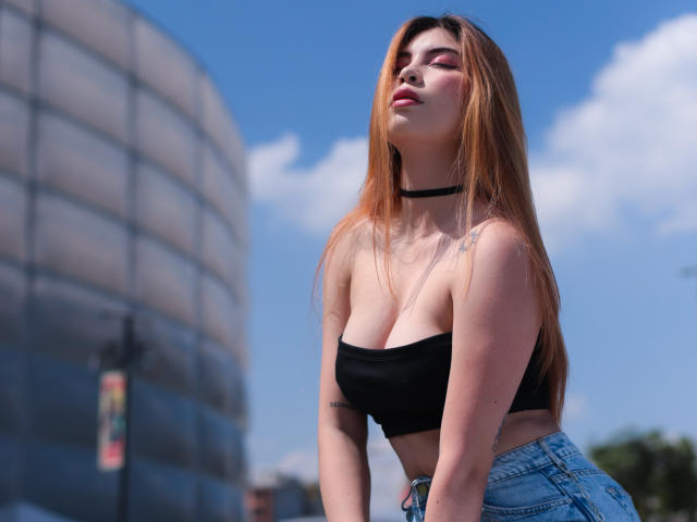 IsaTimeSweet mit langen, orangefarbenen Haaren, einem schwarzen Choker, einem schwarzen Top ohne Träger und blauen Jeans, die ihre großen Brüste und Tattoos zur Schau stellt, vor einem klaren, blauen Himmel..