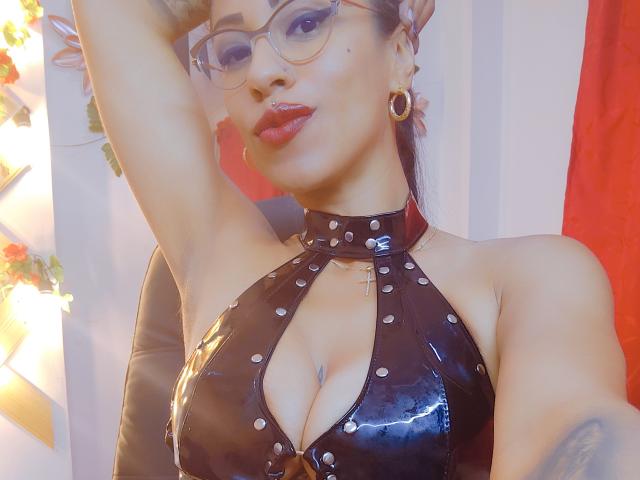 LunaCandy with glasses, red lipstick, black glossy latex halter top, cross necklace, and arm raised, showing her tits and tattoos, against a red and white striped background.