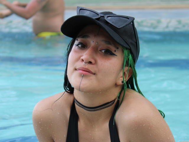 SalomePrince met groen haar, een zwarte muts, een neusring, een lip-piercing en een zwarte choker, waterdruppels op de huid, een zwart bikini, met een zonnebril op haar hoofd, staand in een blauw zwembad..