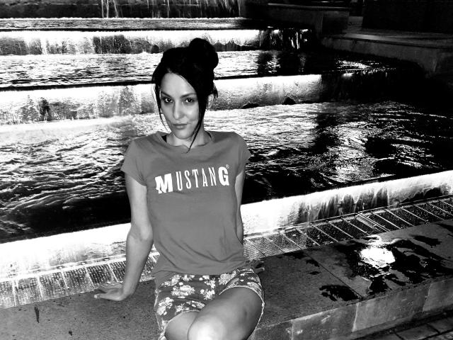 JoliDanielle with dark hair in a bun, wearing a "MUSTANG" t-shirt and floral shorts, sitting by a waterfall, black-and-white, looking at the camera, legs crossed.