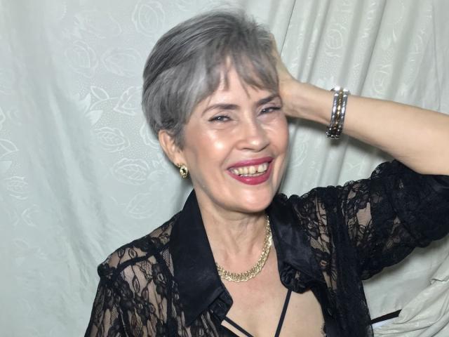 BelleBlondx, short gray hair, smiling with pink lipstick, wearing black lace top, gold necklace, and bracelets, right hand in hair, white background, light skin.