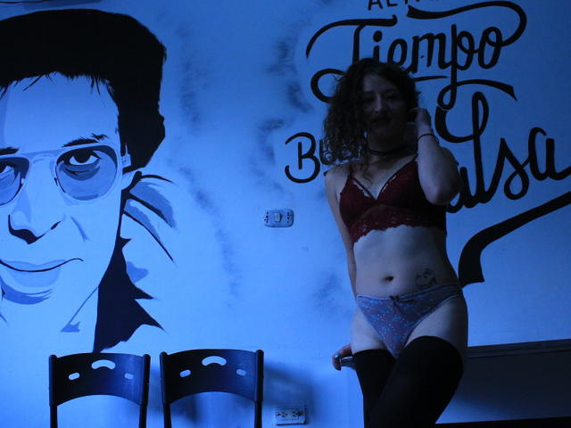 Nataly69Hot está en una habitación iluminada con luz azul, vestida con un sujetador de encaje rojo, ropa interior azul con lunares y medias negras hasta el muslo. Tiene el pelo castaño rizado y un tatuaje en el lado derecho..