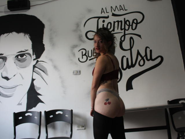 Nataly69, avec ses cheveux bruns bouclés, un soutien-gorge rouge, des culottes grises et des leggings noirs, montre un tatouage de cerises rouges sur sa fesse droite, et se tient à côté d'une chaise noire..