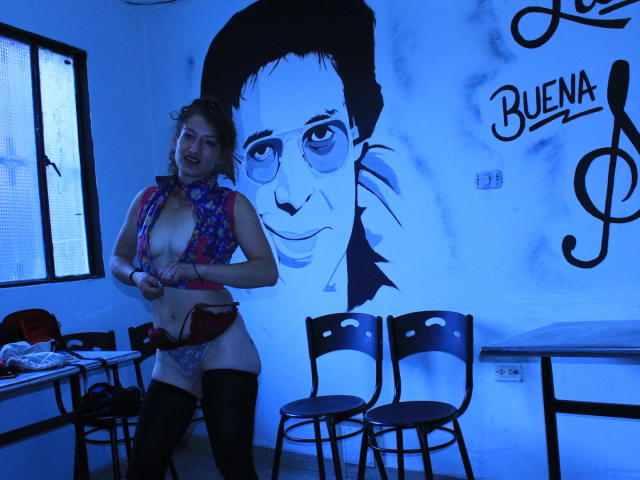 Nataly69, com cabelos castanhos cacheados, vestindo um colete floral vermelho, meias pretas e calcinhas de renda rosa, expondo seus seios em um quarto iluminado por luz azul, com um grande retrato na parede..