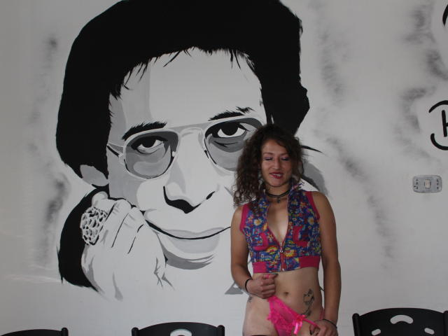 Nataly69Hot, con i capelli ricci, indossa un gilet colorato con motivi floreali, biancheria intima in pizzo rosa e una collana, ed è in piedi di fronte a un murale in bianco e nero raffigurante un uomo con gli occhiali..