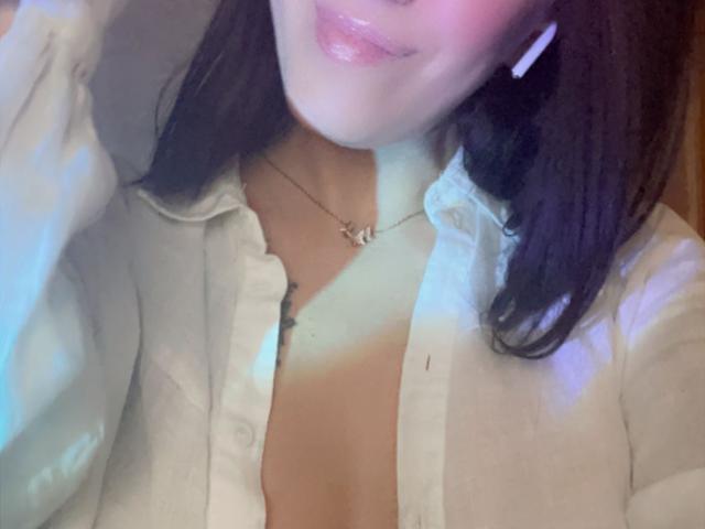 Misstyk with her lips slightly parted, wearing a white button-up shirt, exposing a tan line above her chest, and a delicate necklace, her black hair partially visible.