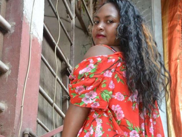 LeCorp with long curly black hair, red floral dress, off-shoulder, looking over shoulder, red lipstick, medium brown skin, standing next to metal bars, confident, sexy, cute.