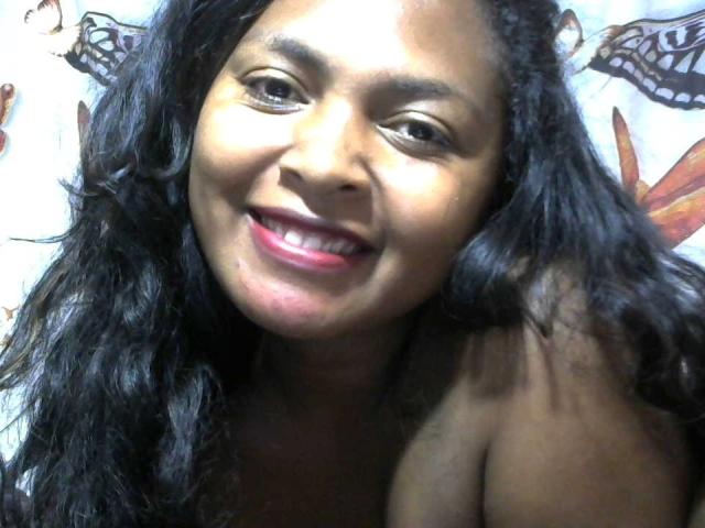 LeCorp's smiling face with dark curly hair, pink lipstick, and bare shoulders, showing her dark brown skin and cute teeth. Her tits are not visible.