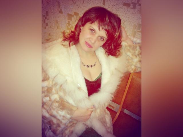 JennyLaFresh, con cabello castaño ondulado, vistiendo un abrigo blanco esponjoso, una blusa negra de encaje y un collar, sonriendo, con las manos en el abrigo, sentada en una silla de madera, con un brillo rosado..