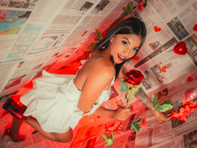Amberv con capelli scuri, rossetto rosso, vestito bianco, che tiene una rosa rossa, guarda indietro, mostrando la schiena e i glutei scoperti, un tatuaggio sulla spalla sinistra, tacchi rossi, fiori rossi..