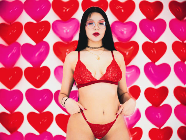 MiaaGrey mit schwarzen Haaren, Brille, rotem Spitzen-BH und -Slips, herzförmigem Hintergrund in Rot und Rosa, Choker-Kette, kleinen Brüsten, flachem Bauch, Hände an den Hüften, selbstbewusst, sexy..