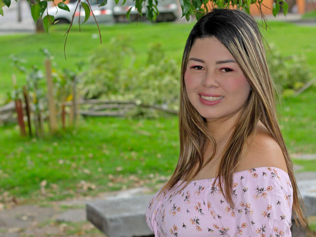 GreeicyCarter com cabelos castanhos longos, blusa rosa com estampa floral e ombro de fora, sorrindo, mostrando os dentes, em pé em uma área gramada, folhas de árvores acima, fundo verde, luz suave, fofa, sexy..