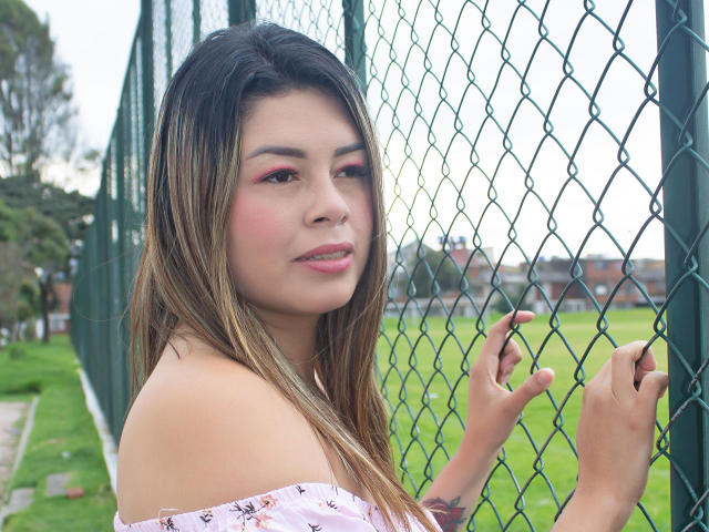 GreeicyCarter, com os seios parcialmente cobertos por uma blusa branca com um ombro descoberto, está em pé perto de uma cerca de arame verde, olhando por cima do ombro, com sombra rosa nos olhos e um leve sorriso..