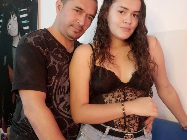 KratosConny, com cabelo castanho ondulado e blusa de renda preta, mostrando os seios, está ao lado de KratosConny, que veste uma camisa preta estampada, e ambas têm os braços ao redor das nádegas uma da outra..