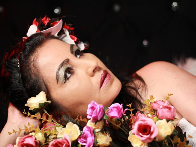 SussyFantasy with dark hair, wearing a flower crown, lying down with pink and yellow roses, smoky green eyeshadow, and shiny pink lipstick, looking up with a seductive gaze.