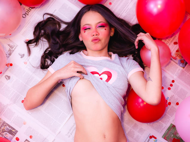 Jazmin Wells, con lunghi capelli neri, ombretto rosa e un tatuaggio a forma di cuore, alza una maglietta grigia per mostrare un ventre piatto e piccoli seni, circondata da palloncini rossi e rosa..
