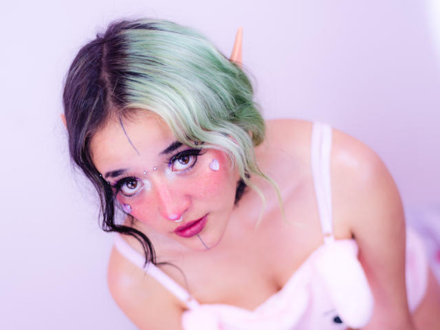 SalomePrince, com cabelo verde e preto, orelhas de elfo, maquiagem em formato de coração rosa, vestido branco e olhos roxos, olhando para cima com os lábios entreabertos, mostrando seus pequenos seios e nariz rosa..