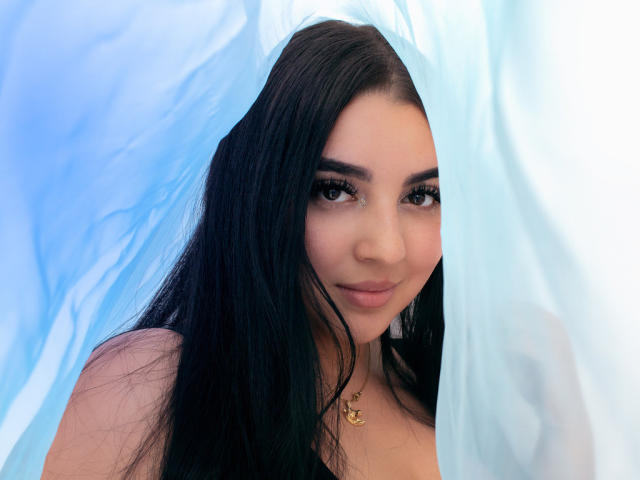 ShinyMurpy, avec ses longs cheveux noirs, ses yeux marrons et son sourire discret, portant un collier en or, entrevue à travers un tissu bleu, révélant son joli visage et ses petites seins..