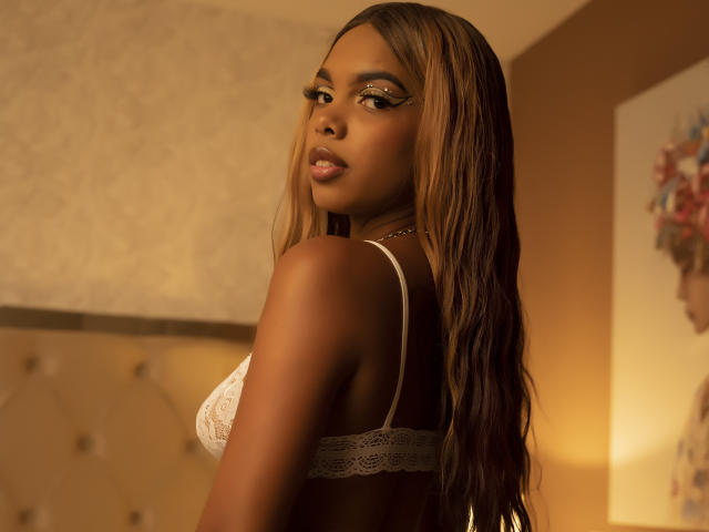 LeyahHill met donkere bruine huid, lang golvend bruin haar, draagt een witte kant bh, kijkt over haar schouder, zachte warme belichting, gouden oogmake-up, subtiele lipstick, in een slaapkamer setting..