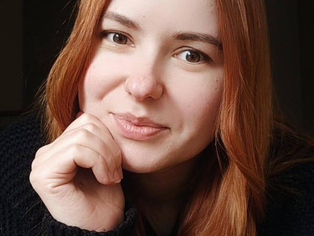 LaylaXDream with red hair, light skin, brown eyes, and a small nose ring, smiling softly, resting her chin on her hand, wearing a black knit sweater.
