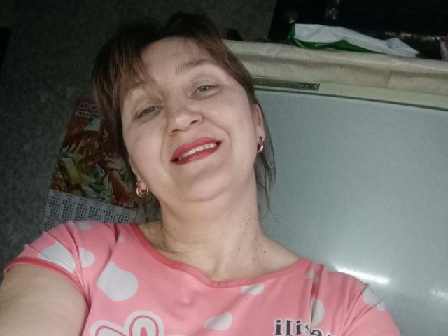 PhebeDi met kort bruin haar, een roze stippen blouse, rode lipstick, lachend, kleine oorbellen, staat voor een wit apparaat. Haar borsten zijn niet zichtbaar..