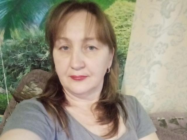PhebeDi ha la pelle chiara, i capelli castani raccolti in due codini, piccoli orecchini, una camicia grigia e rossetto rosso, con un leggero sorriso e un'espressione neutra..