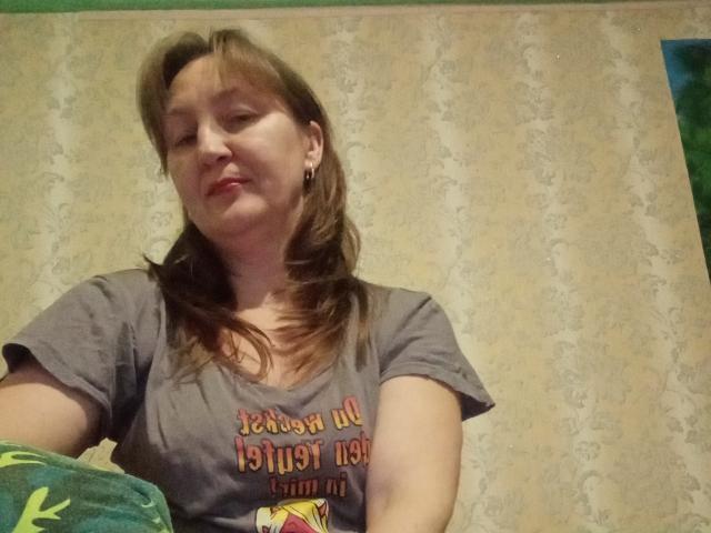 PhebeDi hat braunes, mittellanges Haar, ein graues T-Shirt und ein leichtes Lächeln. Sie sitzt mit einer grünen Decke da und zeigt ihre Brüste. Ihre rosafarbenen Lippen sind entspannt..