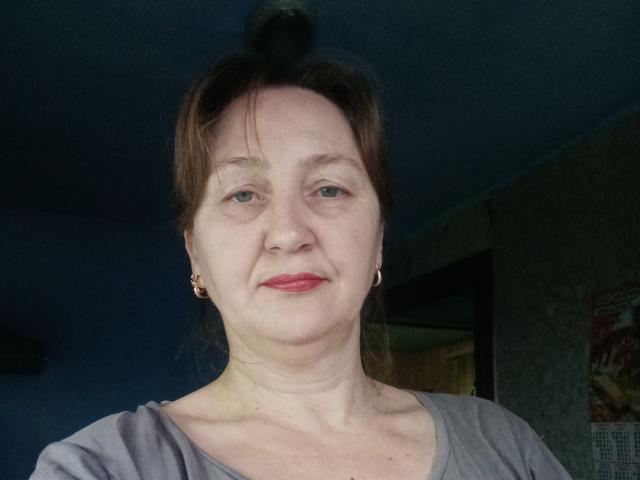 PhebeDi, com pele clara, cabelo castanho preso em um coque desarrumado, brincos pequenos, batom vermelho, camisa cinza e um sorriso discreto, olhando diretamente para a câmera..