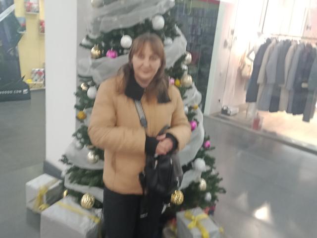 PhebeDi mit braunen Haaren, einer beigen Jacke, schwarzen Hosen und einer schwarzen Tasche, steht vor einem Weihnachtsbaum mit Geschenken, hält die Tasche in der Hand, leicht unscharf..