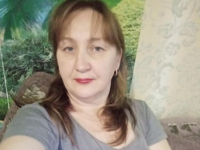 PhebeDi tem a pele clara, cabelo castanho preso em um rabo de cavalo, batom vermelho, pequenas argolas nos ouvidos e uma camisa cinza, com um sorriso discreto, mostrando levemente os seios..