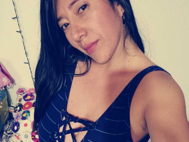 HotPamelaSex with long dark hair, wearing a black striped top with a lace-up front, showing her small tits, smiling softly, standing near colorful, patterned pillows.