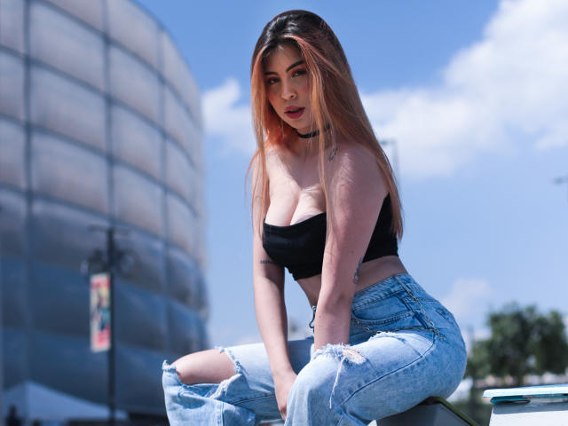 Susan y Jostin, de piel morena y cabello largo y castaño, vestidas con una blusa blanca, inclinadas hacia atrás, mirando hacia atrás. Jostin, con cabello corto y negro y una blusa blanca, agarrándose las nalgas..