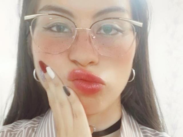 IsaTimeSweet, con cabello negro largo, labios carnosos y una camisa roja de manga larga, tiene las manos cerca de la cara, mostrando sus grandes senos. Lleva jeans azules y está de pie frente a un fondo blanco..
