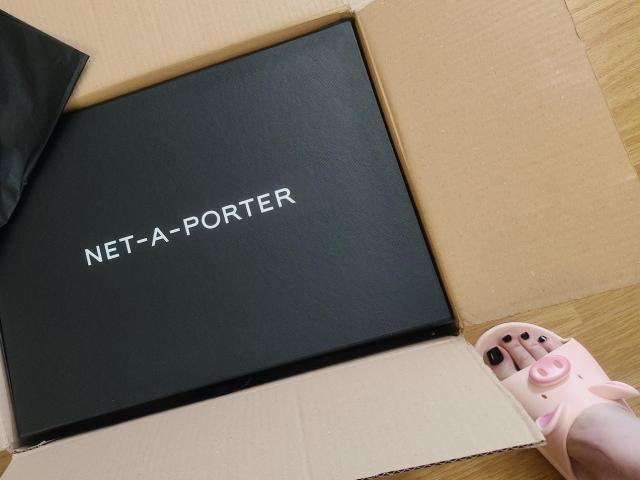 Caixa preta com a etiqueta "NET-A-PORTER", sapatos de bebê rosa com solas pretas, e um pequeno pano preto dentro de uma caixa de papelão bege, tudo sobre uma superfície de madeira..