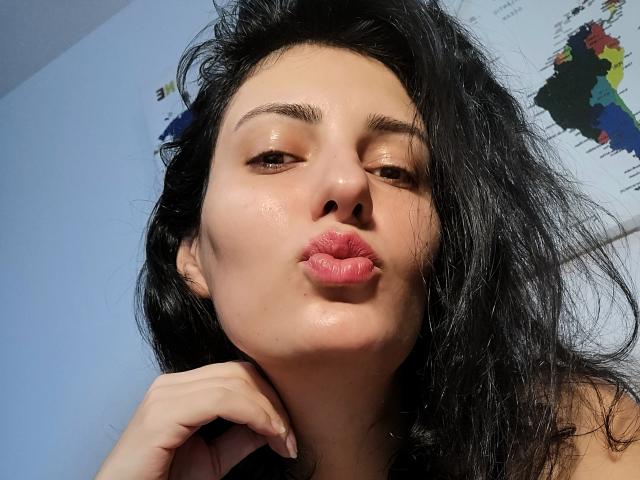 AddictedToUTS has black, messy hair, brown eyes, and is puckering her lips in a kissy face, with shiny skin and a hand near her neck. Her tits are not visible.