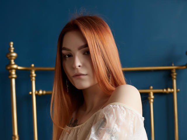 Malena Golden a de longs cheveux roux et lisses, une peau pâle et porte un haut blanc en dentelle avec une épaule dénudée. Elle regarde légèrement sur le côté avec une expression subtile et calme..