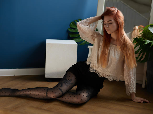 MalenaGolden, avec de longs cheveux roux, un haut en dentelle, des collants noirs à motifs d'étoiles et une jupe noire, assise sur un plancher en bois, une main dans les cheveux, regardant vers le bas, un mur bleu en arrière-plan..