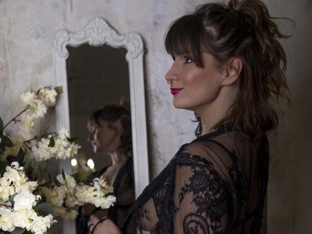 JillianBlack with her tits visible through black lace, nose ring, and red lipstick, reflected in a mirror, standing near white flowers. She has brown hair in a messy ponytail.