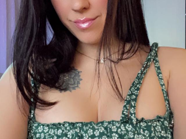 Misstyk with shiny pink lips, dark straight hair, green floral dress, cleavage showing, small tattoo on right chest, slim neck, small silver necklace. Boobs visible.