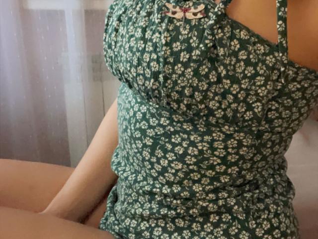 De groene jurk van Misstyk, met witte bloemmotieven, zit strak om haar borsten, waardoor haar rondingen zichtbaar zijn, evenals een klein roze lint aan de linkerkant. Haar armen rusten op haar schoot..