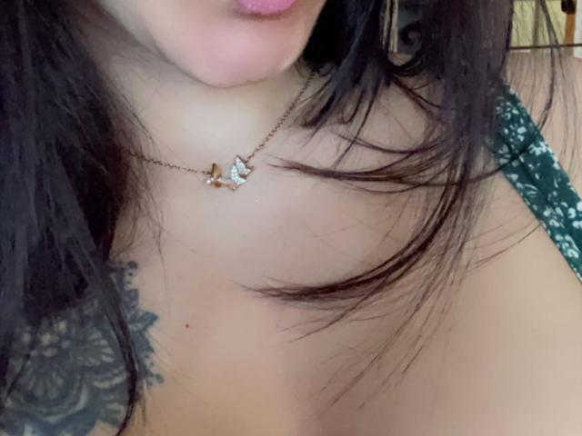 Le labbra rosa e piene di Misstyk, una delicata collana con un piccolo diamante, i capelli neri, una blusa di pizzo verde e un tatuaggio sulla spalla destra. Il seno è visibile nella foto..